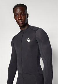 Rashguard noir avec fermeture éclair sur le devant, fabriqué en tissu lisse. Présente un logo blanc sur la poitrine et un design ajusté qui met en valeur les contours du corps.