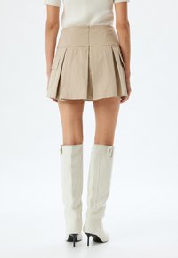 Beige geplooide minirok met zakken en een ritsluiting, gecombineerd met kniehoge witte hakkenlaarzen met gespaccenten.