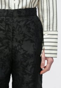 Calças pretas texturizadas com padrão floral, combinadas com uma camisa branca com riscas finas pretas. Vista de perto da cintura e da mão.