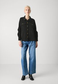 Black corduroy shirt jacket with gold buttons and two chest pockets, paired with light blue wide-leg jeans and black ankle boots.