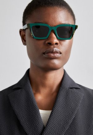 Óculos de sol verdes e estampados com uma armação geométrica; lentes escuras; modelo veste um blazer preto texturizado por cima de uma blusa clara.