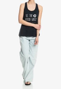 Czarny top na ramiączkach z białym napisem „ALL THE GOOD VIBES” i kwiatowym wzorem; w zestawieniu z jasnoniebieskimi dżinsami o szerokich nogawkach i czarnymi sandałami.
