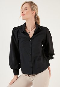 Black button-up blouse with puffed sleeves, made of smooth fabric. Features a classic collar and a curved hemline for a tailored fit.