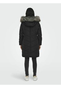 Manteau doudoune noir avec une capuche et une bordure en fausse fourrure. Présente un design matelassé et une coupe mi-longue. Porté sur des leggings gris et des baskets blanches.