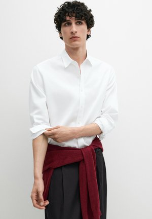 Jonge man die de mouwen van een wit overhemd met knopen oprolt, zwarte broek draagt en een rode trui rond zijn taille heeft gebonden, neutrale achtergrond.