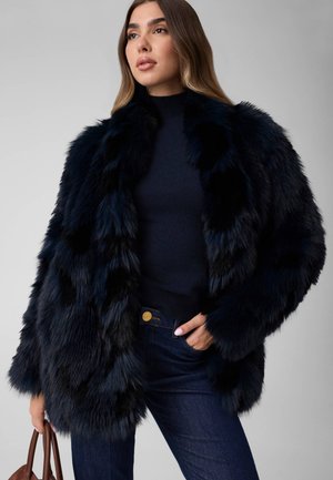Une femme porte un manteau de fourrure noir sur un haut foncé et un jean, et tient un sac à main marron devant un fond de studio gris.