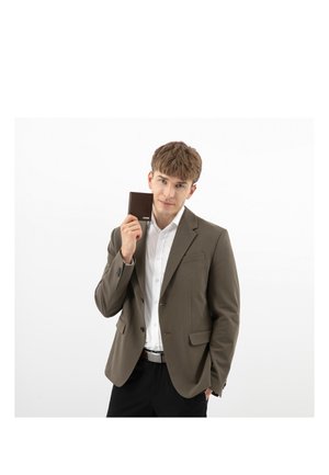 Jeune homme portant un blazer marron et une chemise blanche, tenant un portefeuille marron fermé près de son visage sur un fond blanc uni.