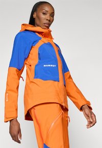 Oranžs un zils jackets ar kapuci, ar rāvējslēdzēja kabatām un "Mammut" logo. Audums izskatās teksturēts un izturīgs.