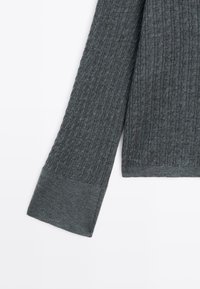 Pull en maille gris foncé à manches longues avec un motif torsadé et de larges poignets côtelés sur fond blanc.