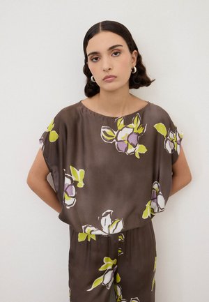 Vrouw met kort donker haar draagt een bruine bloemenprint top en bijpassende broek, staat met handen achter de rug tegen een effen witte achtergrond.
