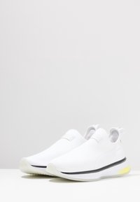 Białe sneakersy wsuwane z teksturowanej tkaniny, gładkim, zaokrąglonym noskiem oraz kontrastującą czarną podeszwą z żółtym akcentem na pięcie.