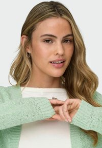 Mint green cardigan with ribbed cuffs, worn over a white textured top. The knit features an open design and relaxed fit.