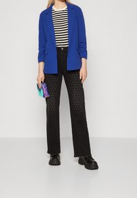 Blazer blu su una camicia a righe bianco e nera, abbinato a pantaloni neri strutturati con motivi a pois e scarpe nere massicce.