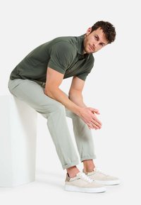 Korte mouwen olijfgroen polo, lichtbeige chino's en crèmekleurige sneakers met textuuraccenten, zittend op een witte pedestal.