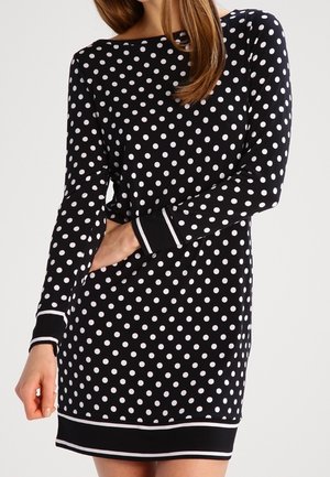 Femme portant une robe noire à manches longues avec des pois blancs, des poignets rayés blancs et un ourlet, debout devant un fond uni.