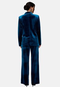 Samtanzug in tiefem Blaugrün mit einem maßgeschneiderten Blazer und weit geschnittenen Hosen. Verleiht eine glatte Textur und dezenten Glanz. Rückansicht gezeigt.