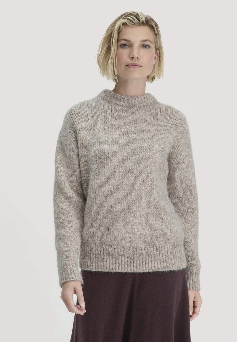 Blonde Frau, die einen strukturierten beigefarbenen Strickpullover und einen dunklen Rock trägt, steht vor einem schlichten hellen Hintergrund.