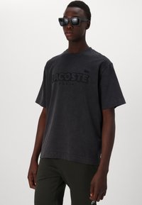 Čierne oversized tričko s textúrovaným logom "LACOSTE", vyrobené z bavlny. V kombinácii s tmavozelenými nohavicami a čiernymi slnečnými okuliarmi.