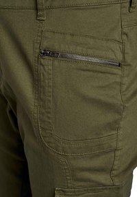 Olivu krāsa kravas bikses ar struktūrētu audumu, ar kabatu ar rāvējslēdzēju sānos un pastiprinātu šuvju detaļām.