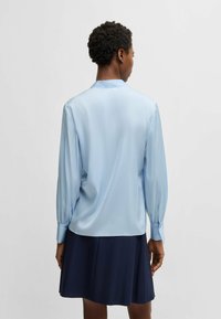 Blusa di raso azzurro chiaro con maniche lunghe a palloncino, polsini con bottoni e colletto. Il retro mostra una texture liscia e lucida.