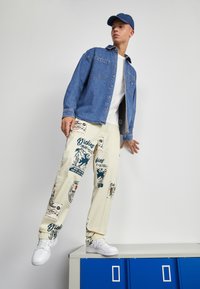 Μπλε πουκάμισο τζιν με κουμπιά, λευκό μπλουζάκι, κρεμ παντελόνι με εκτυπώ�σεις μπλε γραφικών και λευκά αθλητικά παπούτσια με μπλε καπέλο. Στέκεται μπροστά σε μπλε ντουλάπι.