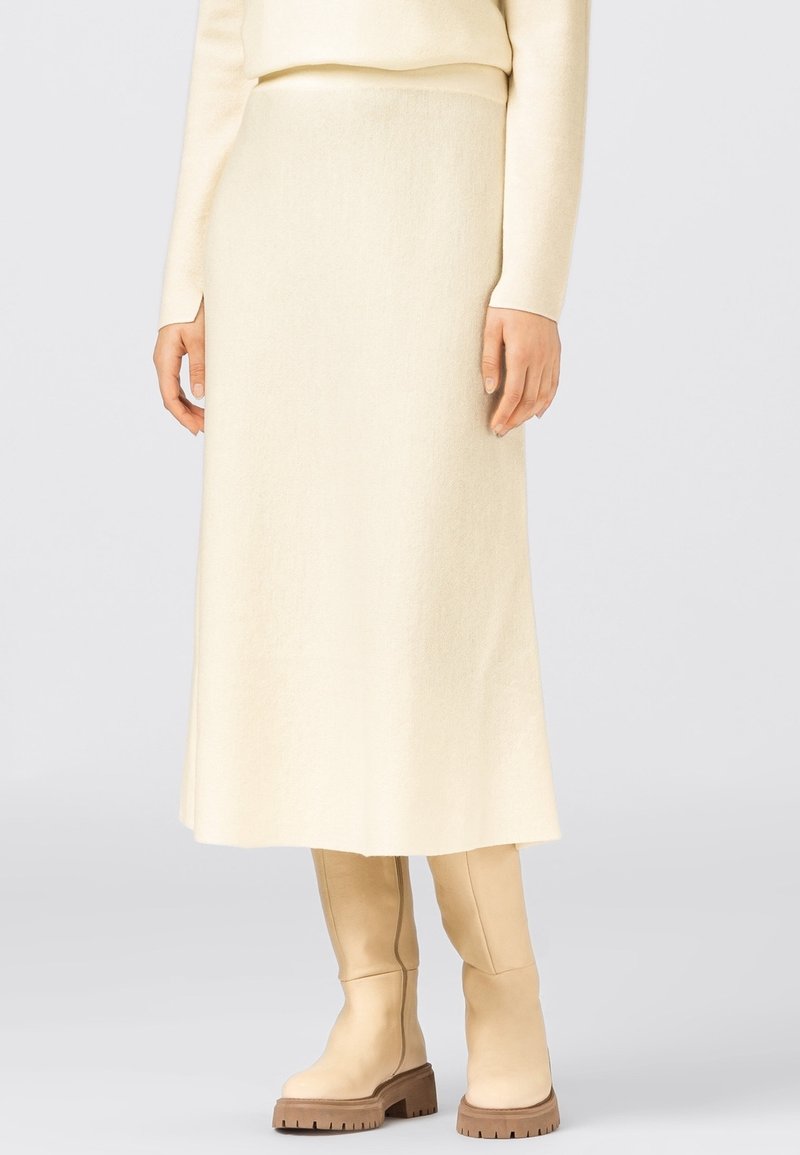 Jupe mi-longue en tricot de couleur crème, avec une texture lisse, une forme évasée et une taille haute. Associée à des bottes beige montant jusqu'aux genoux.