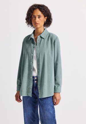 Frau mit lockigem Haar, die ein hellgrünes, strukturiertes Hemd mit Knöpfen über einem weißen Oberteil und blauen Jeans trägt, und vor einem schlichten weißen Hintergrund steht.