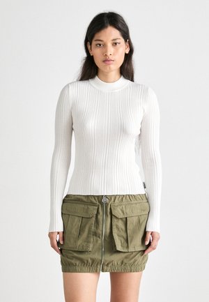 Haut à manches longues blanc côtelé associé à une jupe utilitaire verte, dotée d'une fermeture éclair à l'avant et de grandes poches. Design simple et ajusté avec un tissu texturé.