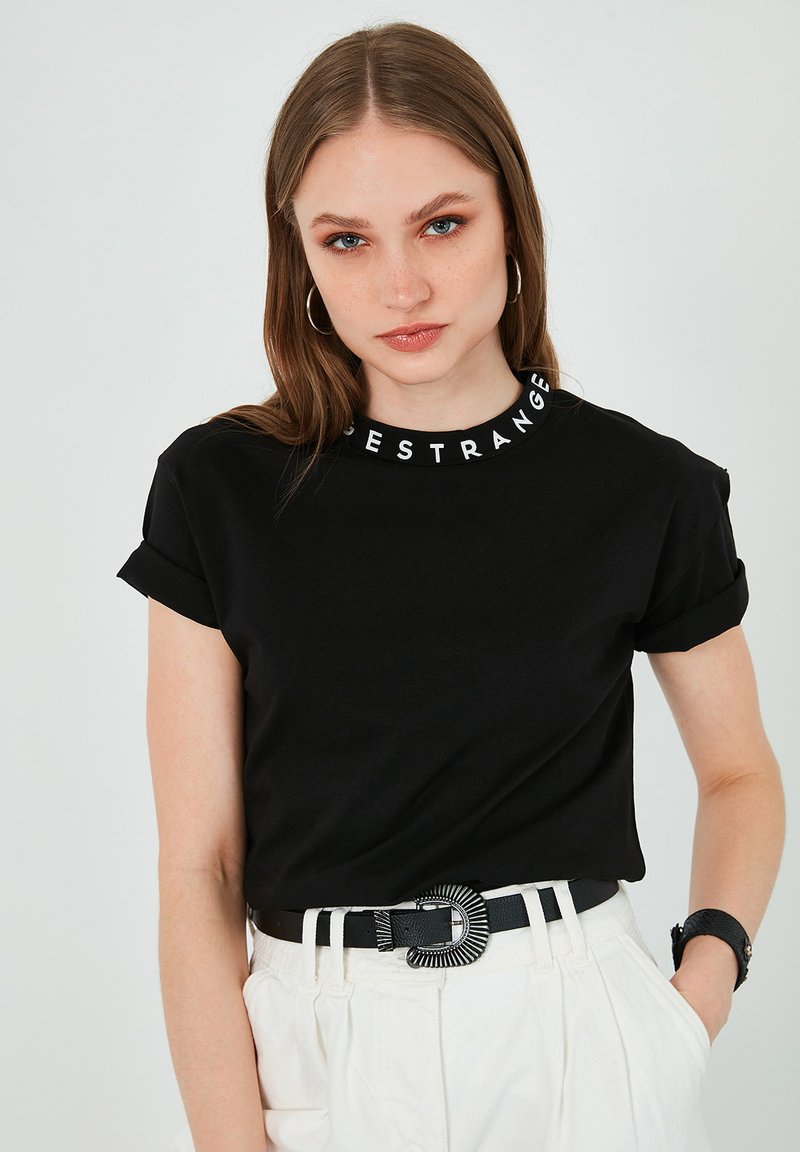 Must tee-shirt, mille äärtega, millel on valge tekst "STRANGE" krael. Kandub musta vööga ja heledate pükstega.