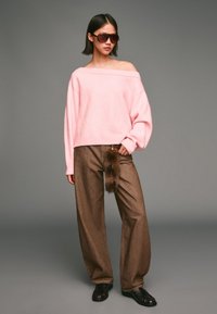 Hellrose Off-Shoulder-Pullover, braune weite Hose mit einem Kunstfell-Akzent und dunkelbraune Slipper. Sonnenbrille rundet den Look ab.