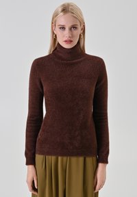 Maglione marrone a collo alto con trama, maniche lunghe; materiale morbido e peloso; forma aderente. Indossato con gonna verde a pieghe.