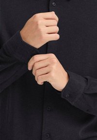 Camicia nera con colletto button-down, maniche lunghe e bottoni in contrasto. Il tessuto sembra testurizzato con una leggera lucentezza.