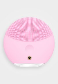 Dispositif de nettoyage facial en silicone rose avec une surface texturée de petites brosses rondes. Forme circulaire avec une base blanche et un connecteur doré.