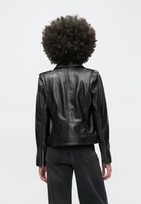 Göndör hajú személy fekete bőrkabátot és sötét farmert visel, hátulról látható, egyszínű háttér előtt.