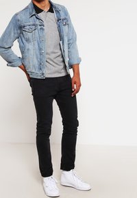 Veste en jean à délavage bleu, polo gris, jean noir et baskets blanches. Dotée d'une fermeture à boutons et d'une coupe régulière.