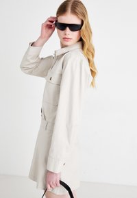 Lys beige skjortekjole med krage, lange ermer, knappedetaljer og innsnevret midje. Båret med sorte solbriller og holder en svart veske.