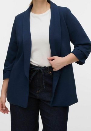 Kobieta w granatowej, otwartej marynarce z podwiniętymi rękawami, białej koszulce i ciemnych, wysokich dżinsach z paskiem przewiązanym w pasie.