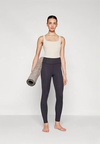 Šviesiai smėlio spalvos liemenė ir juodi aukšto liemens tamprės, su tekstūruota jogos kilimėliu, susuktas po viena ranka, stovint ant paprasto fono.