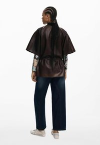 Top oversize in pelle marrone con maniche corte, stretto in vita, indossato sopra una camicia a maniche lunghe con stampa e abbinato a jeans blu scuro.