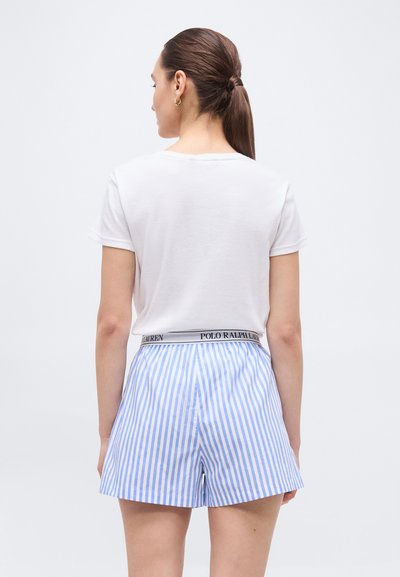 Γυναίκα με καστανά μαλλιά σε αλογοουρά, φοράει λευκό κοντομάνικο πουκάμισο και μπλε-λευκά ριγέ σορτς με ζώνη Polo Ralph Lauren.