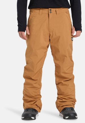 Pantalons de snowboard marron avec une texture lisse, fermeture à double bouton et deux poches sur le devant, portés avec des boots noires.