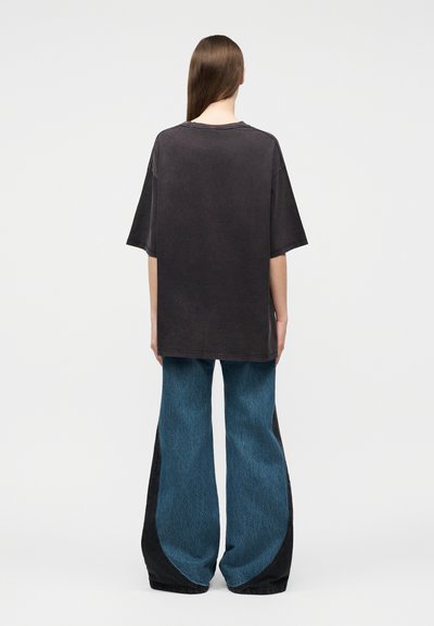 Άτομο με μακριά μαλλιά φοράει υπερμεγέθη μαύρο T-shirt και φαρδιά μπλε τζιν με μαύρες πλαϊνές λεπτομέρειες, στέκεται πλάτη σε μονόχρωμο φόντο.