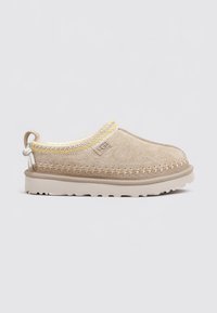 Chaussure UGG en suède beige, à enfiler, avec semelle épaisse, bordure cousue jaune et beige, et languette à l'arrière, présentée de profil sur fond blanc.