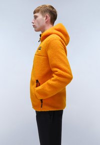 Orange fleecejacka med huva, texturerad yta, framfickor med dragkedja och sidfickor. Dragkedjesmycken i svart. Buren av en modell.