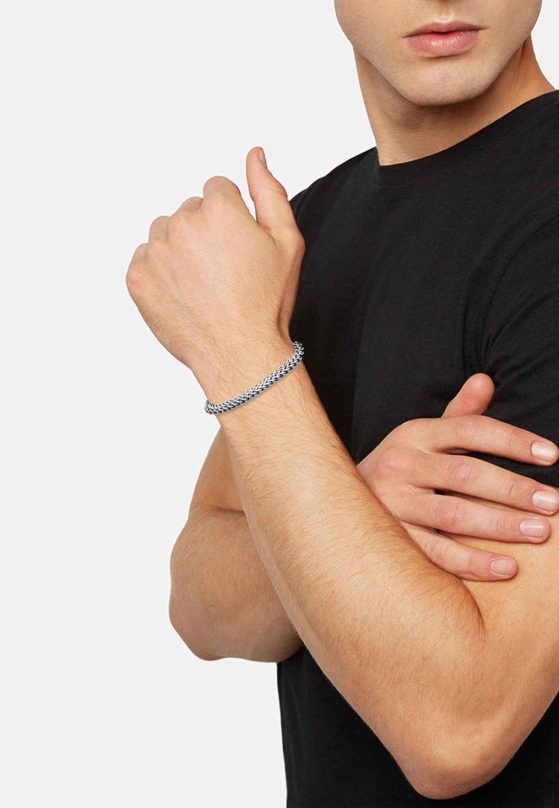 Braccialetto in metallo argento con un design intrecciato, indossato su un polso. La persona indossa una maglietta nera, con le braccia incrociate e uno sfondo neutro.