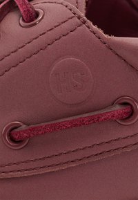 Close-up van een bordeauxleren schoen met gestikte naden, bordeauxsuede veters door ronde oogjes geregen, en het geperste "HS"-logo.