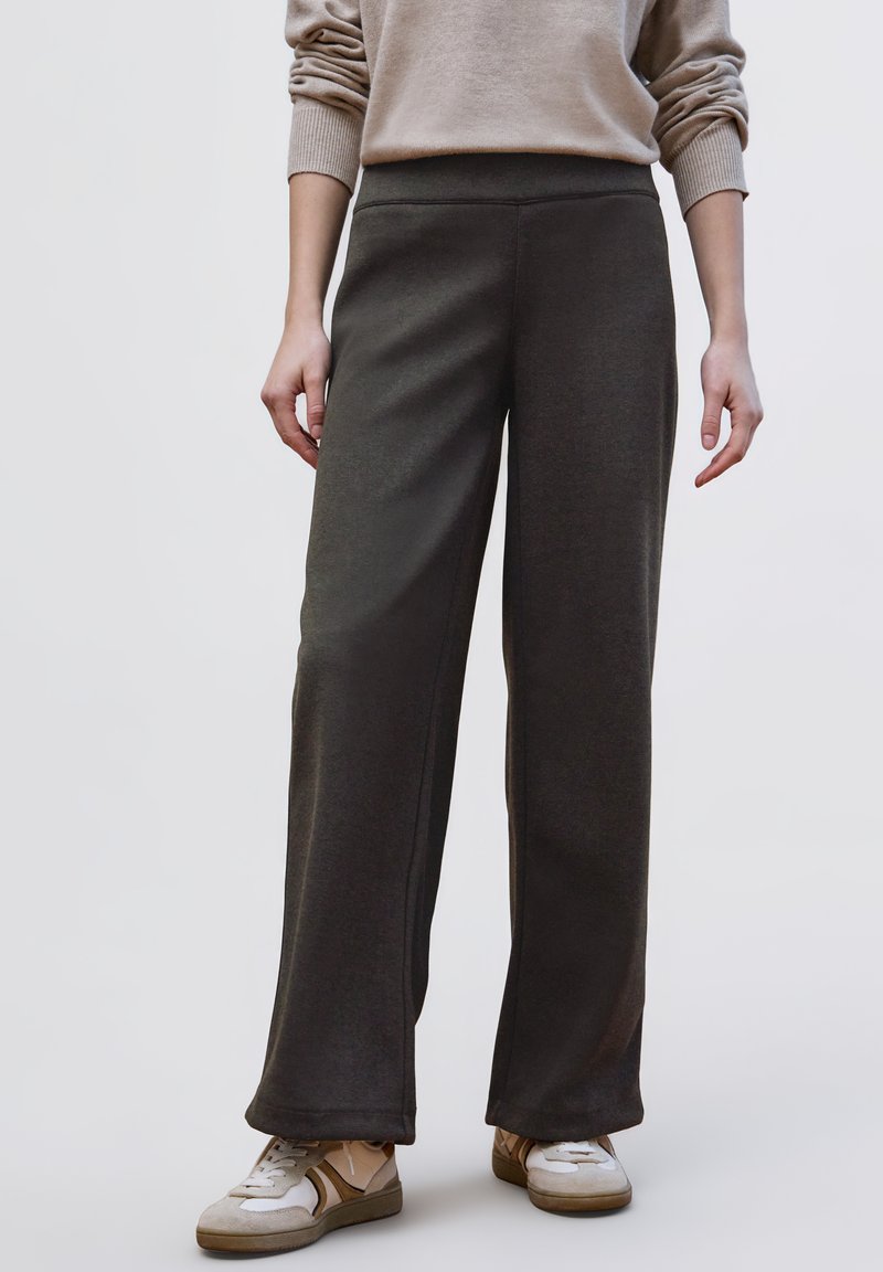 Tmavě šedé široké kalhoty z měkké látky s vysokým pasem a minimem švů, doplněné světlými teniskami.