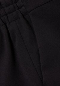 Černá látka s hladkým povrchem a drobným leskem. Detail pružného pásku se sklady, nediferencovaný vzor a žádné viditelné kovové části.