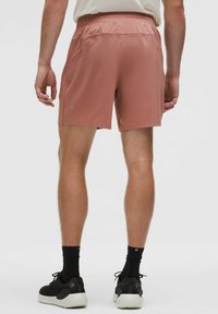 Férfi sportnadrág, világos rózsaszín, könnyű anyagból készült, gumis derekú, két oldalsó zsebbel, fekete sportzoknikkal és cipőkkel kontrasztban.