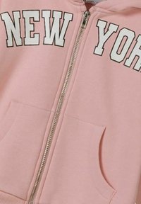 Veste à capuche zippée rose en tissu doux, avec un grand texte blanc "NEW YORK" sur la poitrine et deux poches avant.